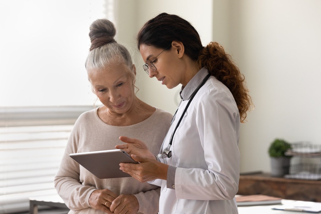 Serious GP doctor showing tablet screen to old 70s female patient, explaining electronic prescription, medical screening, examination result, giving consultation. Woman visiting practitioner