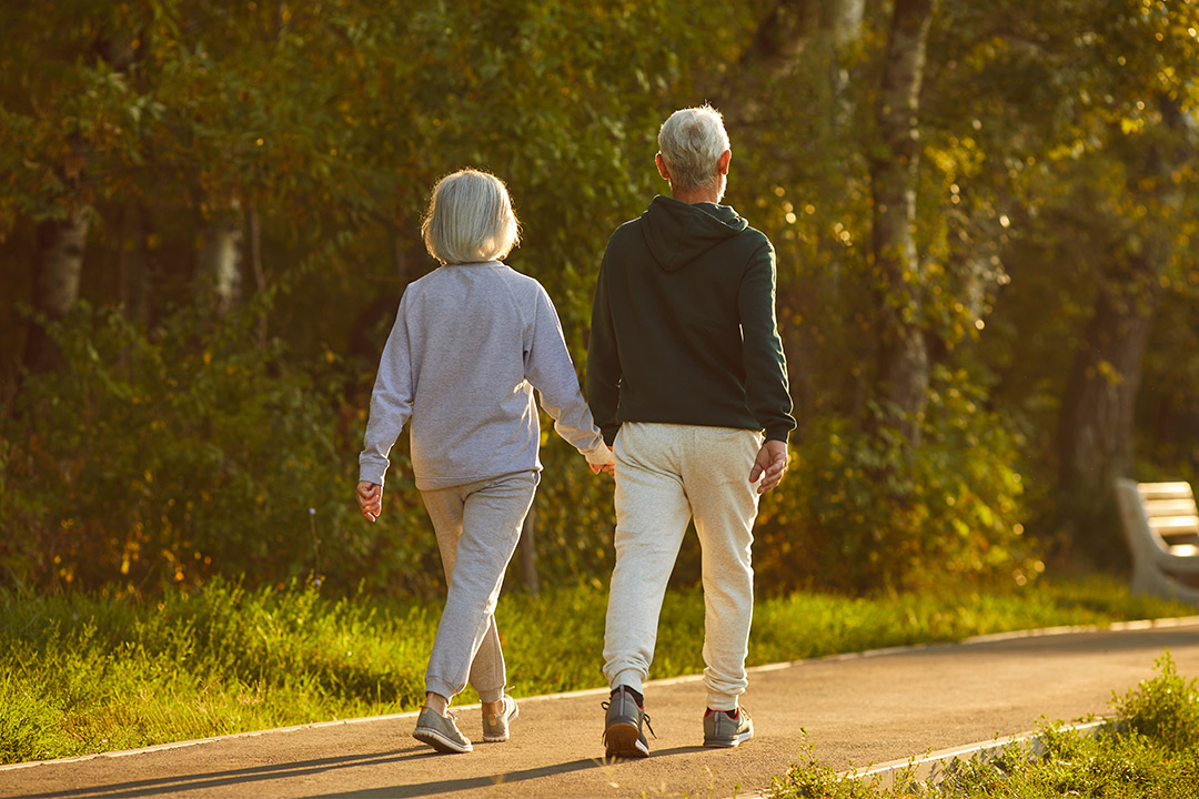 An older couple strolls hand-in-hand along a park path, surrounded by greenery and trees.
