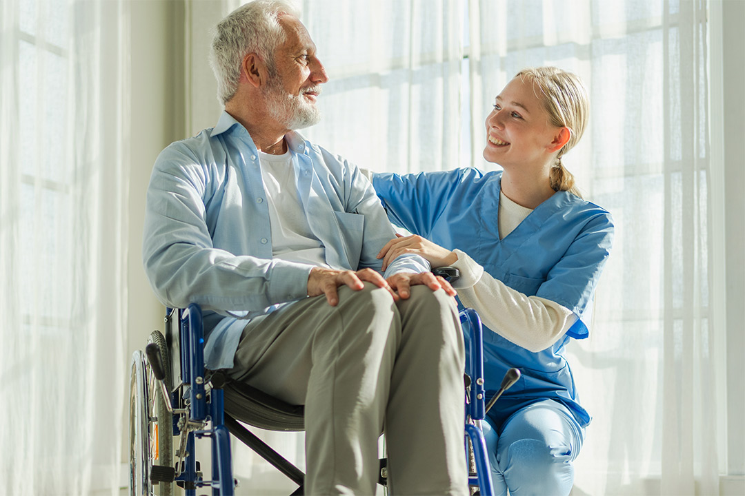 A healthcare worker in scrubs supports an elderly man in a wheelchair, emphasizing skilled nursing in chronic illness care.